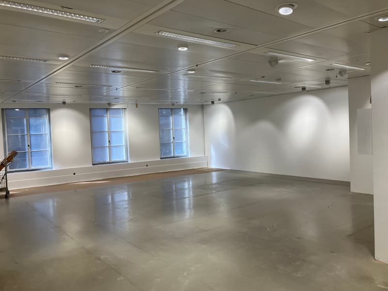 Spacious, empty room with concrete floor and white walls, three windows, track lighting, and a cart.