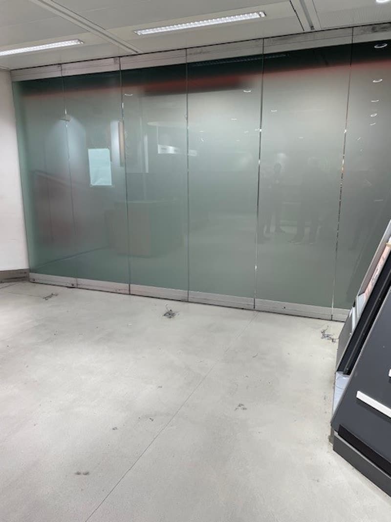 Empty room with large glass wall, light grey floor, shadows on the wall, two people reflected in the glass.