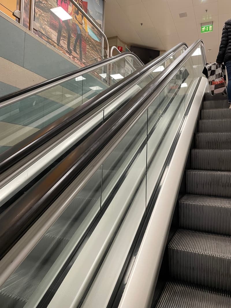 Two escalators going up and down, modern glass design, metal handrails, and a person on the right using the stairs