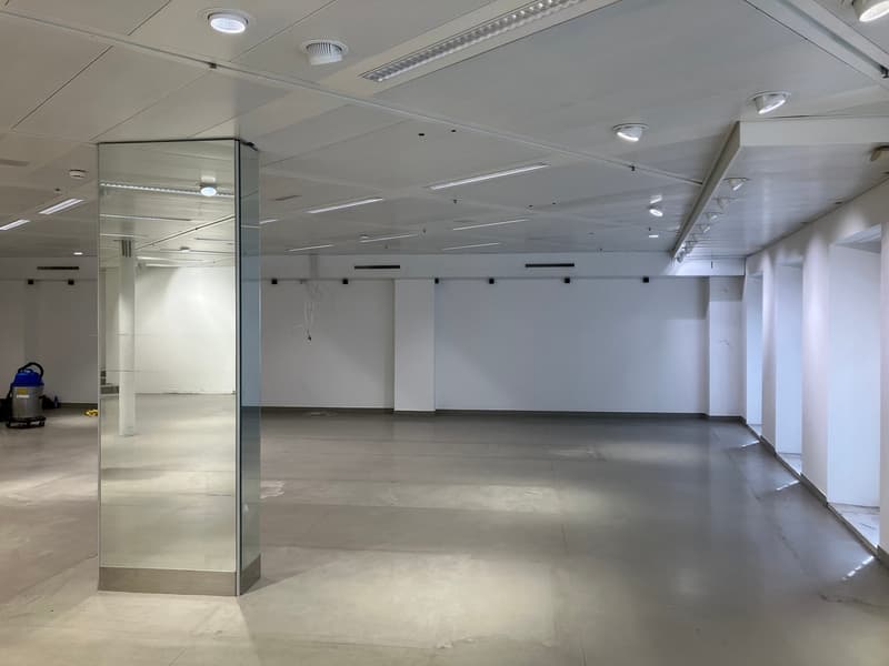 Empty, large room with tiled floors, white walls, high ceilings, and recessed ceiling lights. A glass partition runs down the middle of the room. There are some electrical and plumbing fixtures visible on the walls.