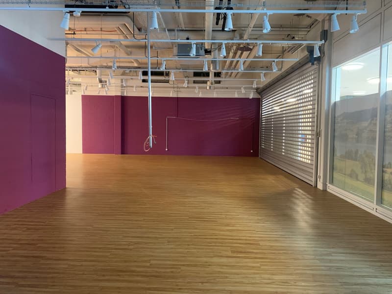Large open space with hardwood floors, burgundy-colored walls, and an exposed ceiling with ventilation ducts and lighting fixtures. The space has large windows along one wall, providing natural light. The room appears to be a commercial or office space, p