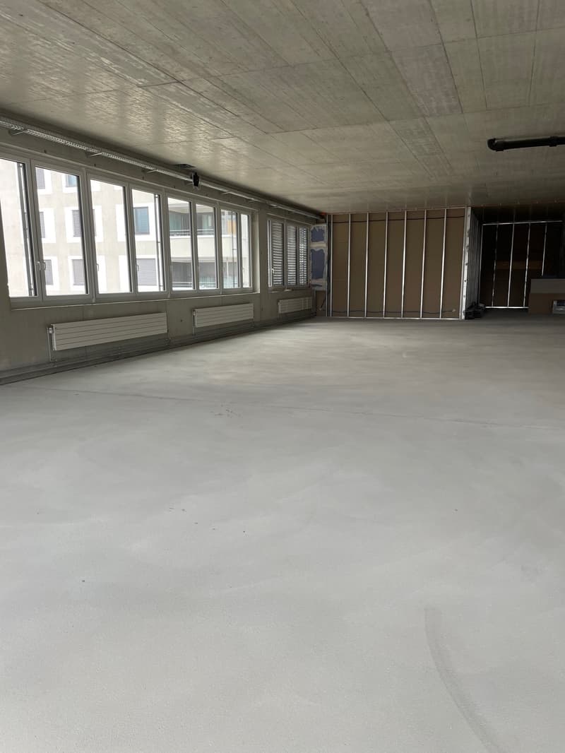 Empty room with concrete floor, large windows with shutters, wall mounted heaters, beams on ceiling, unfinished partition wall, entrance on right