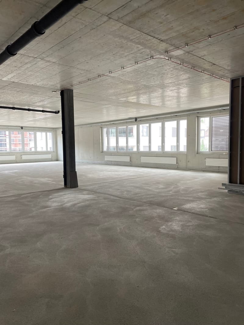 Empty office space with high ceiling, concrete floor, large windows, central pillar, white walls, beams in the ceiling, and large windows.