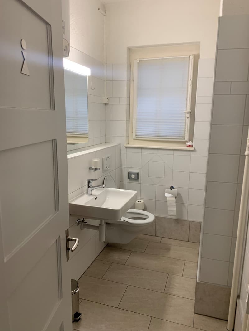 white tiled walls, white toilet, white sink, small window with blinds, toilet paper holder, mirror with light, trash can