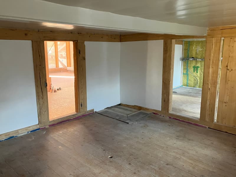 This image shows an unfinished room with exposed wooden beams and a wooden floor. The room appears to be in the process of renovation, with some construction materials visible on the floor. The room has large windows that provide natural light, and there