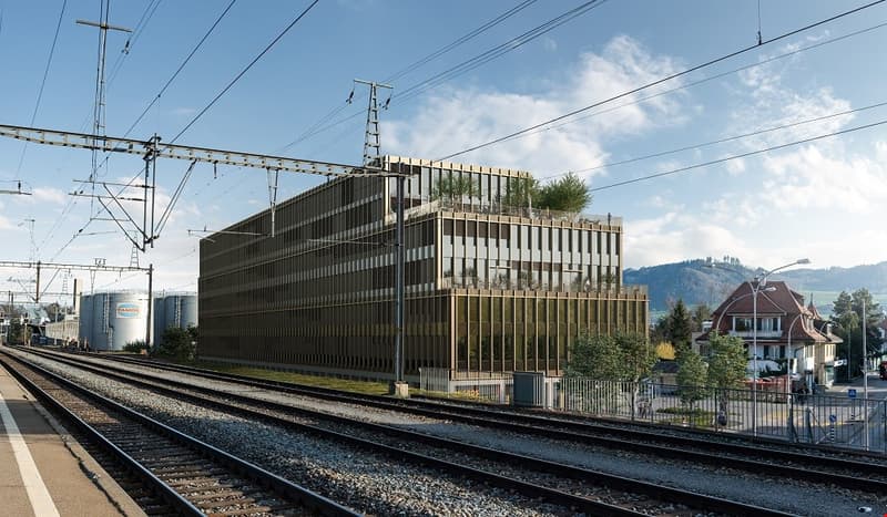 Modern office building with glass facade, located near a railway track, surrounded by trees and a house