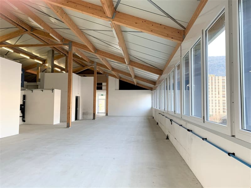 The image shows a large open commercial space with a high wooden ceiling and exposed beams. The space has concrete flooring and large windows along one wall, providing natural light. The space appears to be unfinished or under renovation, with no furnitur