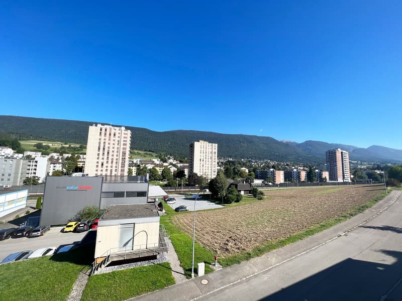 The image shows a cityscape with several high-rise apartment buildings and a large open field in the foreground. The buildings have a modern, residential appearance and are surrounded by trees and greenery. The sky is a clear, bright blue, indicating a su