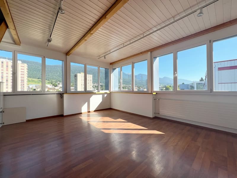 Spacious room with large windows offering a scenic view of the surrounding mountains and cityscape. The room has a wooden floor and a paneled ceiling with exposed wooden beams. The room appears to be well-lit with natural light streaming in through the wi