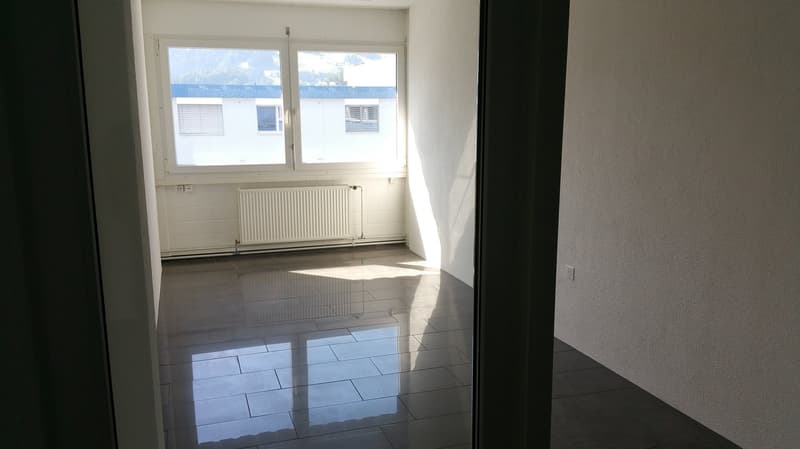 Empty room with white walls, large windows, and tiled flooring. The room appears to be a living room or bedroom with a radiator visible on the wall.