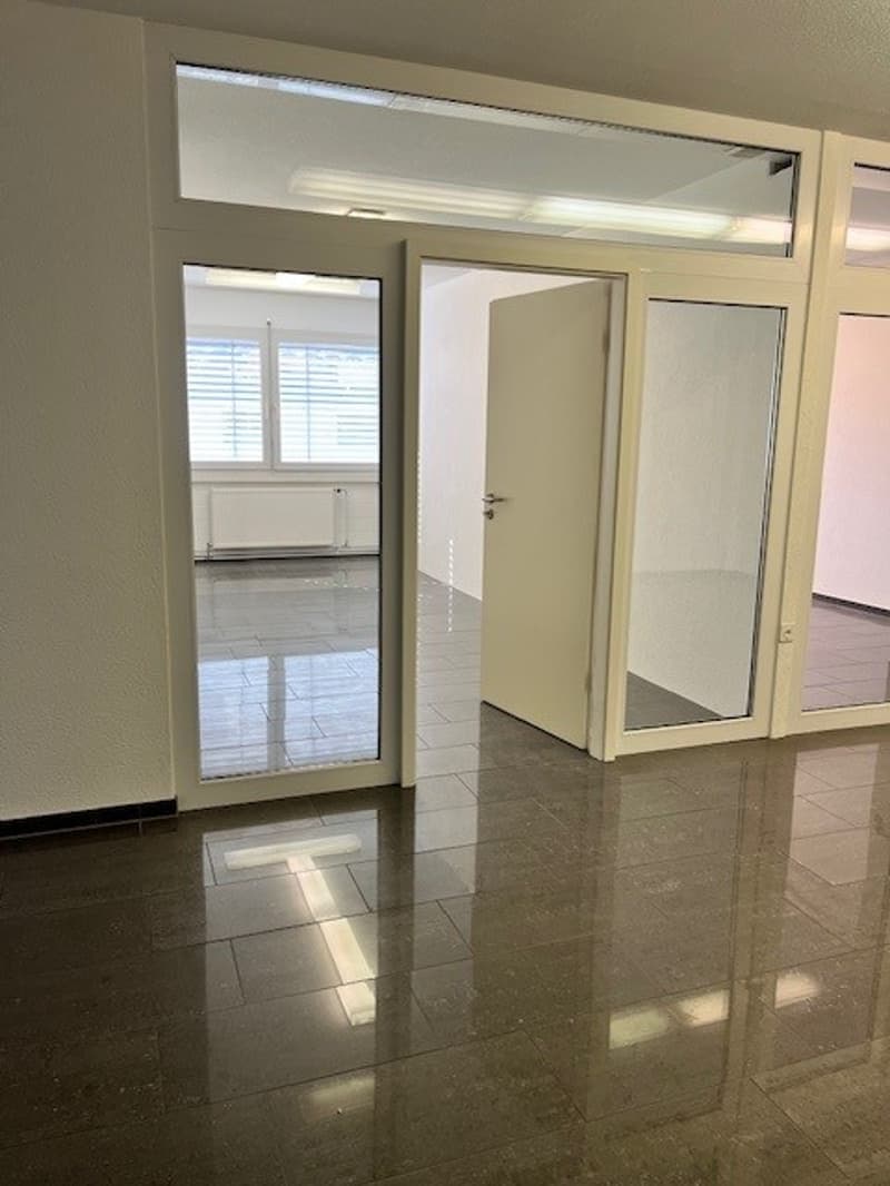 This image shows an empty room with a tiled floor, white walls, and large windows. The room appears to be in a residential or commercial building, with a door visible leading to another room or area.