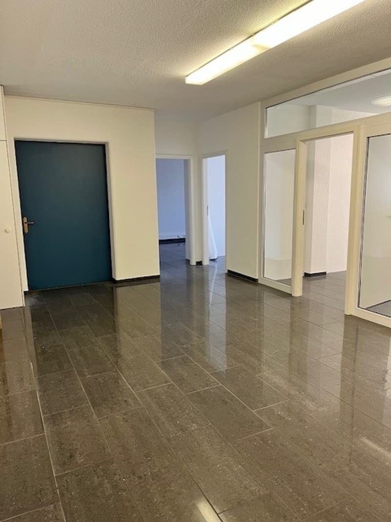 The image shows a large, open office space with a tiled floor and white walls. There are several doors visible, including a blue door in the foreground. The space appears to be well-lit with overhead lighting.
