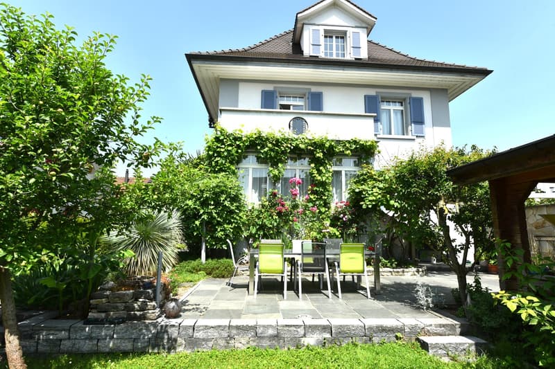 Two-story house, white facade, brown roof, blue shutters, balcony with vine covering, dining table, chairs, garden with plants and flowers