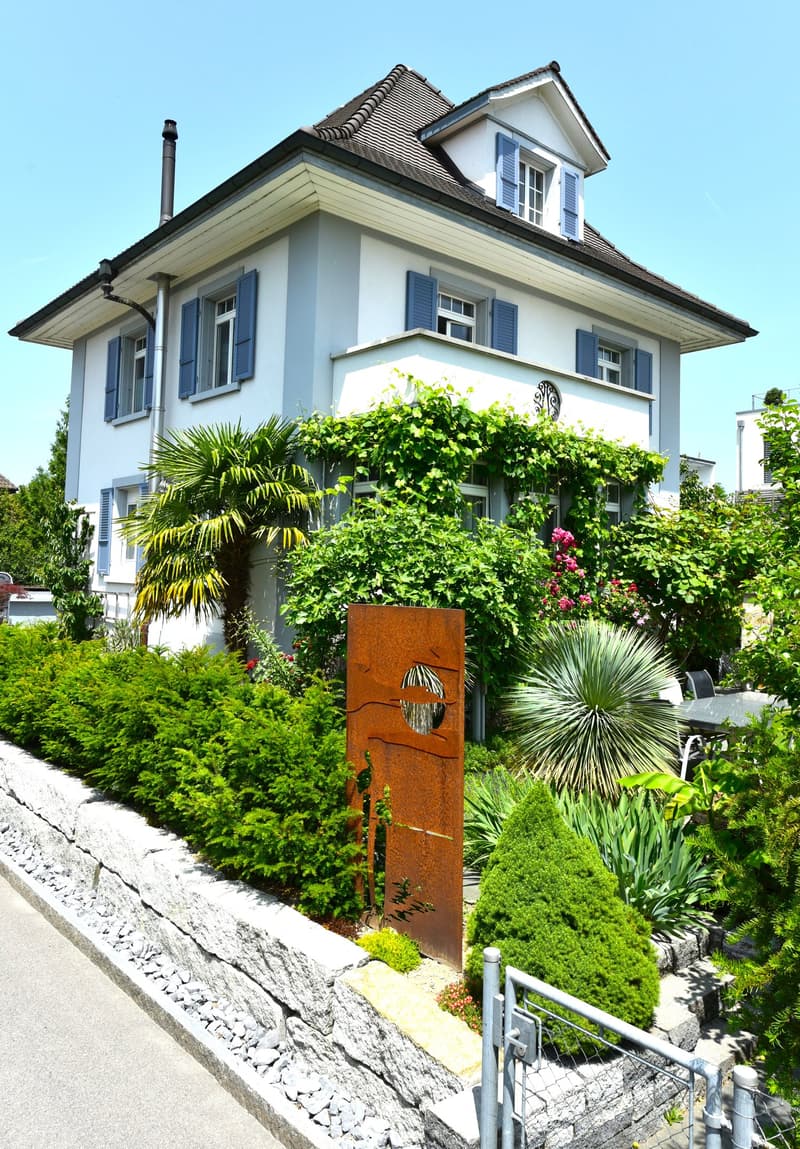 Raised ground floor, 2 story house, white exterior, brown roof, garden with a table and chairs, outdoor seating area