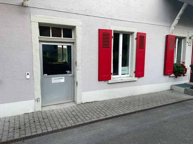 A white-walled building with three windows and a door, all with red shutters. The building is on the street side with a brick pathway.