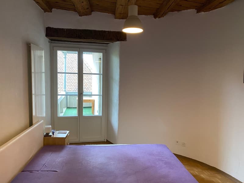 bedroom with wooden floor, white walls, bed, bedside table, sliding glass door to balcony, wooden ceiling