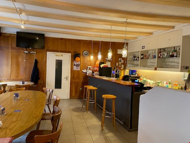 The image shows the interior of a restaurant or bar. It has a wooden ceiling with exposed beams, a long wooden table, and a bar area with stools. There is a TV mounted on the wall and various shelves and cabinets behind the bar, suggesting it is a fully e