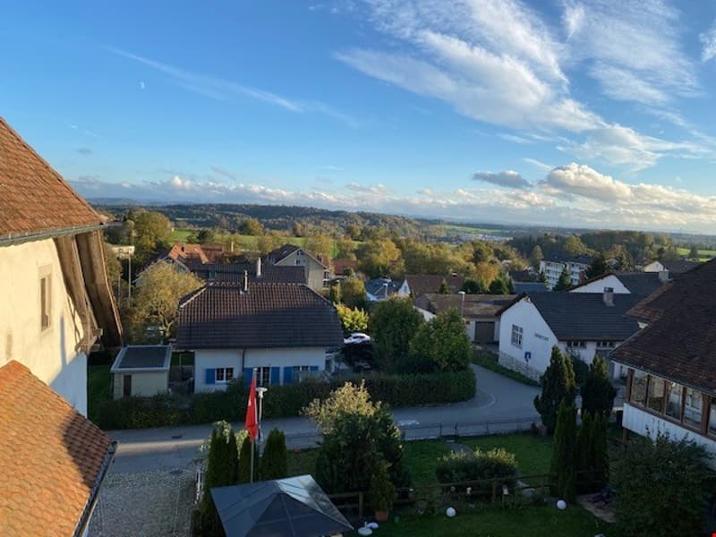 The image shows a scenic view of a small town or village nestled in a hilly, rural landscape. The town is surrounded by trees and greenery, with a mix of residential and commercial buildings visible. The sky is clear with some wispy clouds, creating a pic