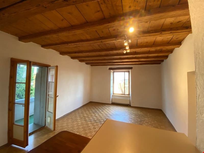 Spacious room with wooden ceiling beams, white walls, and a large window. The room has a tiled floor with a checkered pattern.