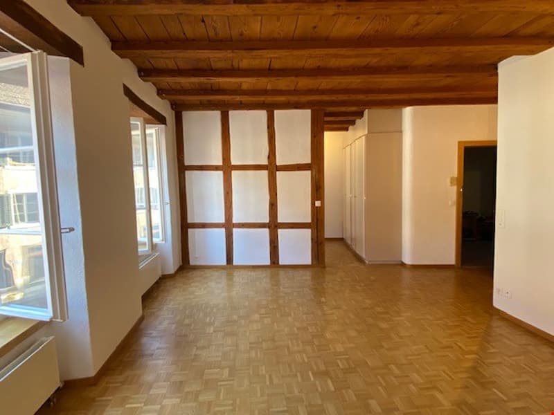 Large open space with wooden beamed ceiling, white walls, and hardwood flooring. The room has a set of large windows and a built-in cabinet or closet.