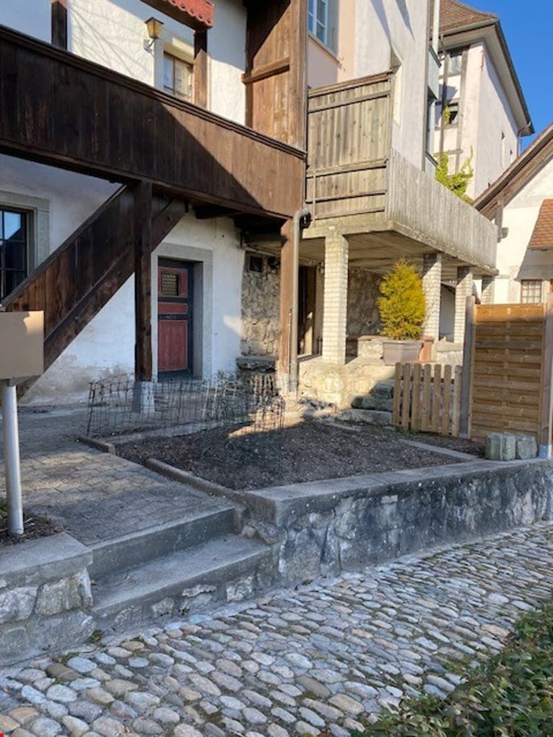 Two-story house with a wooden exterior, stone foundation, and a covered porch. The house has a wooden balcony and a cobblestone walkway leading to the entrance.