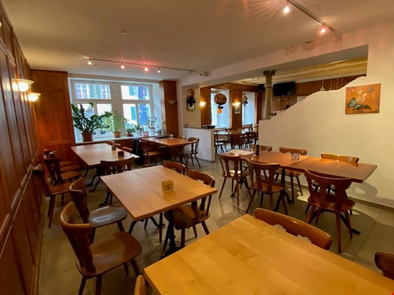 The image shows the interior of a restaurant or cafe. The space has wooden tables and chairs, with a warm and cozy atmosphere. The walls are decorated with artwork and there are string lights hanging from the ceiling, creating a pleasant ambiance.