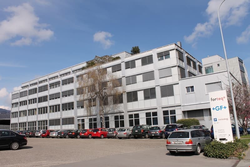 Multi-story office building with a large parking lot in front, located in an urban area with a blue sky and some clouds in the background.