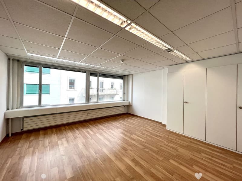 An empty room with wooden floors and white walls. Large windows with white window sill and green shutters. White built-in cabinets and a white ceiling with integrated fluorescent lighting.