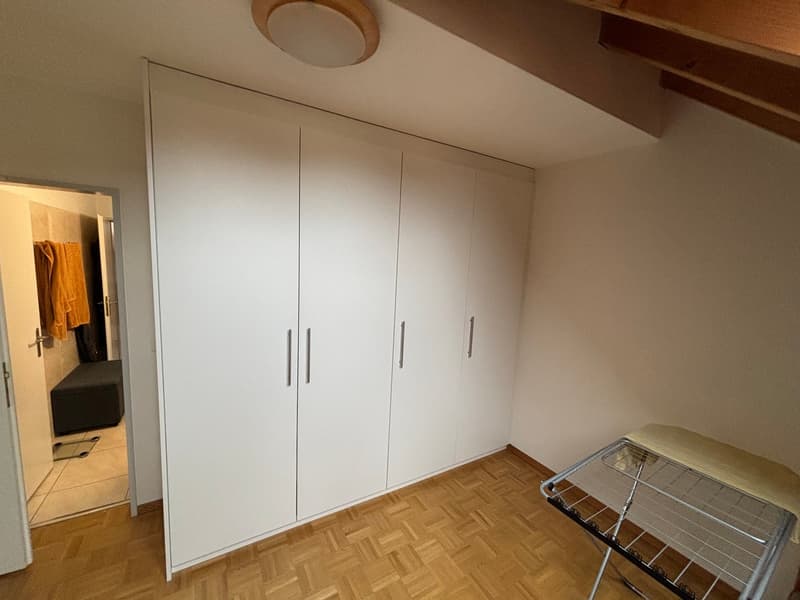 A room with a large white wardrobe, a wooden floor, and a white ceiling with a ceiling lamp. There is a door leading to another room with a towel hanging on the wall.