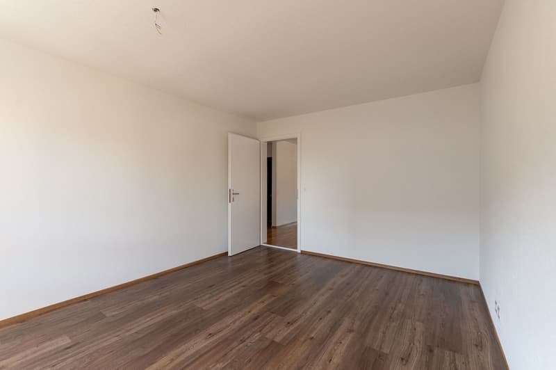 Empty room with white walls, wooden floor, open door leading to another room, power outlet on the wall