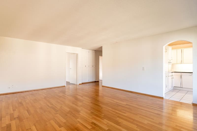 empty room, wooden floor, white walls, power outlets, open door to kitchen, kitchen with dishwasher and cabinets