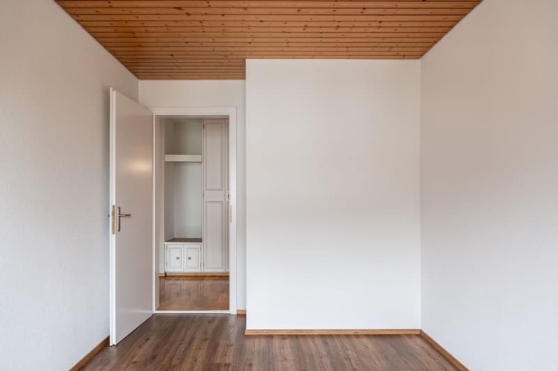 Empty room with white walls, wooden floor, open door, closet with shelves, small bench