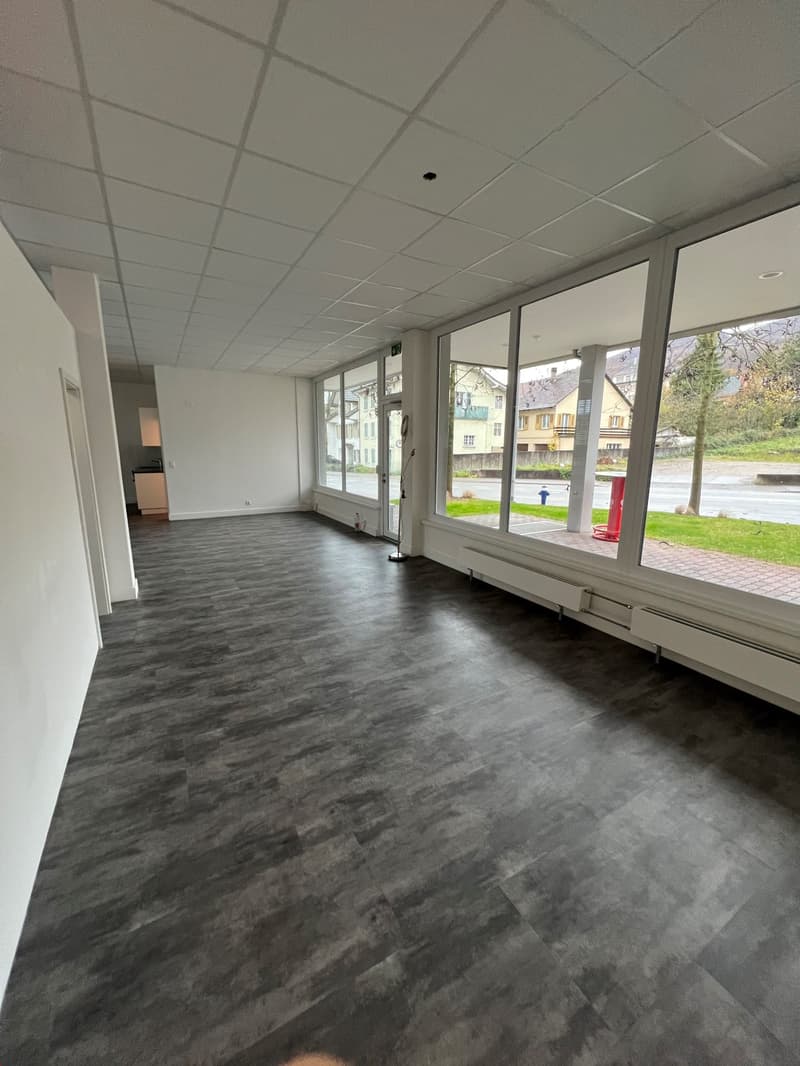 The image shows a large, open office space with a tiled ceiling and large windows overlooking a grassy outdoor area. The floor is covered in a dark gray, textured vinyl or laminate flooring. The space appears to be empty and ready for use as an office or