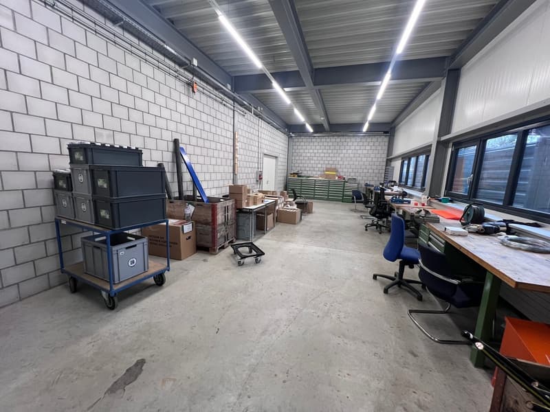 Industrial room with white brick walls, fluorescent lighting, multiple storage boxes, chairs, desks, and scattered items