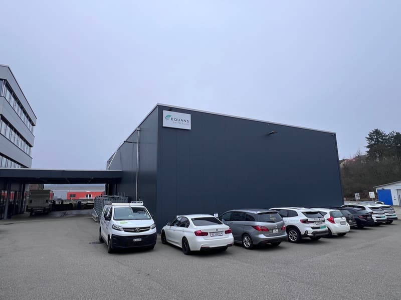 Large building with logo of Equans Switzerland, parking lot with several cars, grey building facade, gray parking lot.