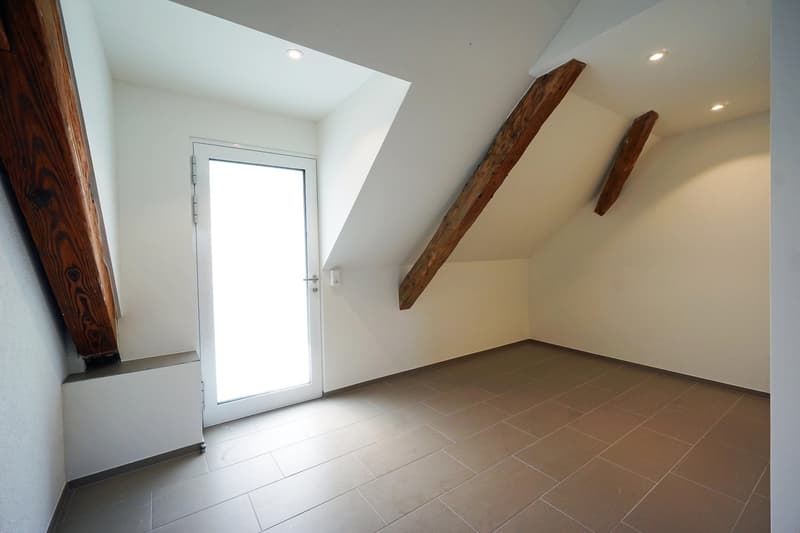 Spacious room with white walls, wooden beams, and a glass door leading to a balcony or terrace. The room has a tiled floor and recessed lighting.