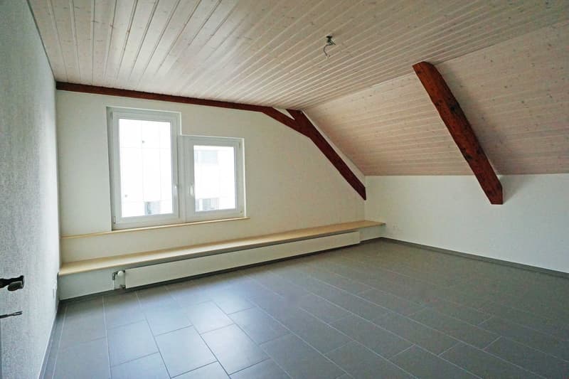 Attic room with slanted ceiling, white walls, wooden beams, and large windows. The room has a tiled floor and a built-in bench or shelf along one wall.