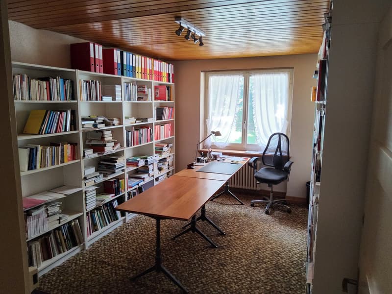 Wooden desks, black office chair, bookshelves filled with books, lamps, large window with curtains, radiator, brown carpet