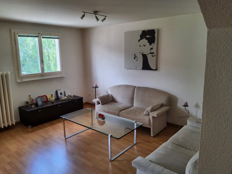 white walls, wooden floor, glass window with view of trees, glass table, couch, beige couch, black coffee table, artwork, decorative items