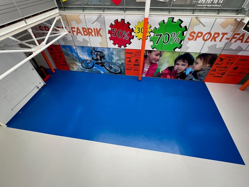 The image shows the interior of a retail store, with a large blue surface in the foreground and various promotional signs and displays on the walls. The store appears to be focused on sports and outdoor equipment, with signs advertising discounts and sale