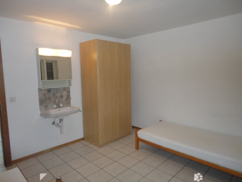 Small room with white tiled floor, white single bed, wooden wardrobe, sink with mirror, power outlet