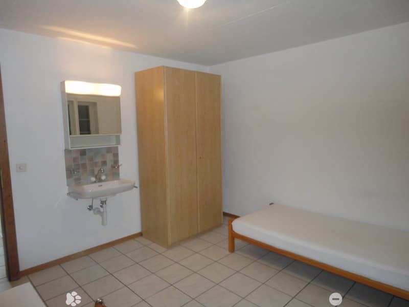 Small room with white tiled floor, white single bed, wooden wardrobe, sink with mirror, power outlet