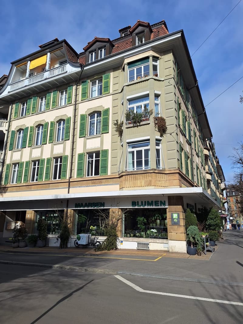 A multi-story building with green shutters, a balcony on the second floor, and a sign that reads 'Maarsen Blumen' at the front.