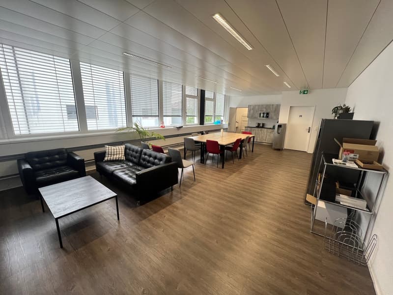 The image shows a modern and spacious office space with a large open floor plan. The room features a comfortable seating area with a black leather sofa and coffee table, as well as a dining table with red chairs for meetings or collaborative work. The spa