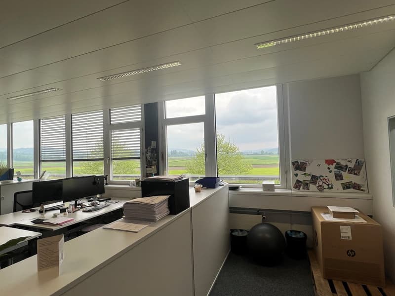 Spacious office with large windows overlooking a scenic outdoor landscape, desks with computers and office equipment, shelves and storage units, and a bulletin board with photos and documents.