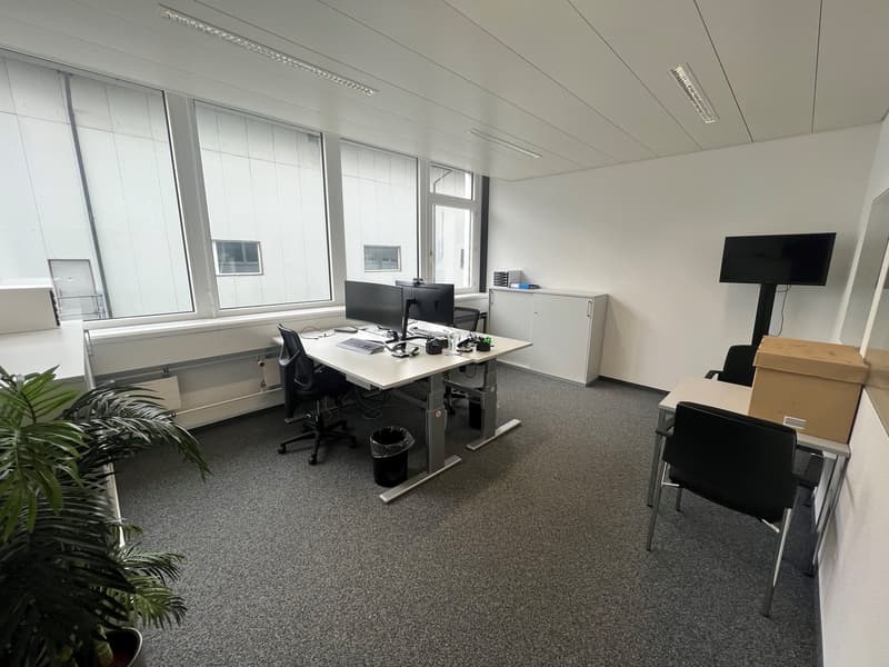 The image shows a modern office space with a large desk, computer setup, and storage cabinets. The room has large windows providing natural light, and there is a plant in the corner. The floor is covered in gray carpeting, and the overall atmosphere appea