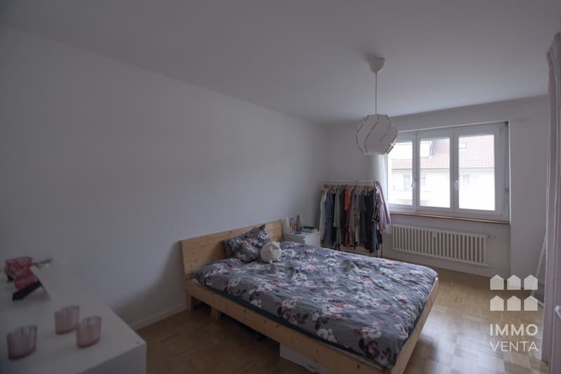 white walls, wooden floor, a bed with floral cover, night stand with lamp, window, radiator, hanging clothes, white desk