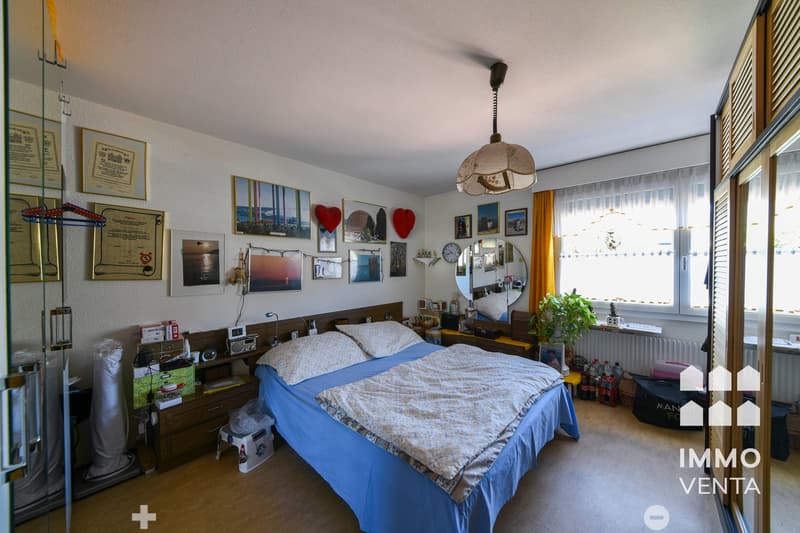 Bedroom with bed, wooden headboard, pillows, blanket, nightstands, lamps, framed pictures, mirror, plants, radiator, window with curtains, wooden floor.