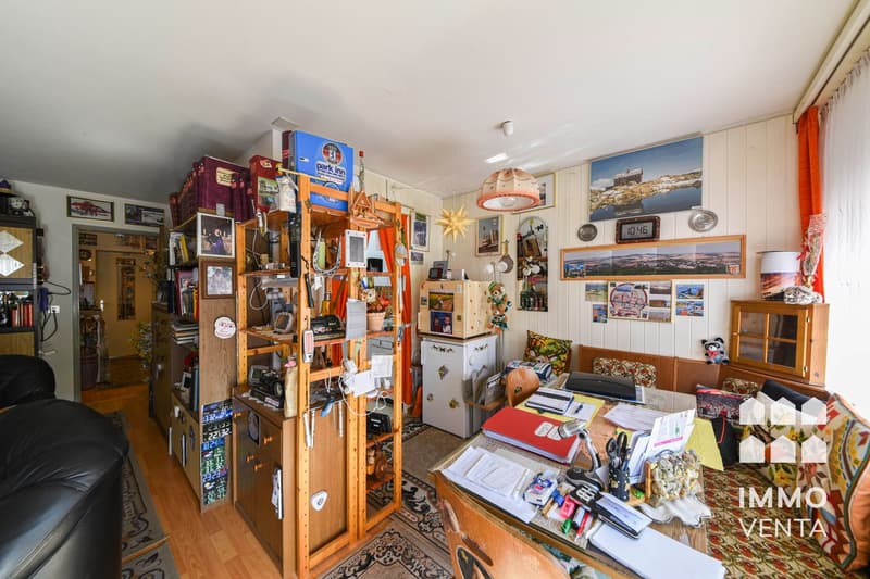 cluttered room with wooden furniture, shelves, books, and various items, a desk with papers and objects, a couch, and a black leather chair