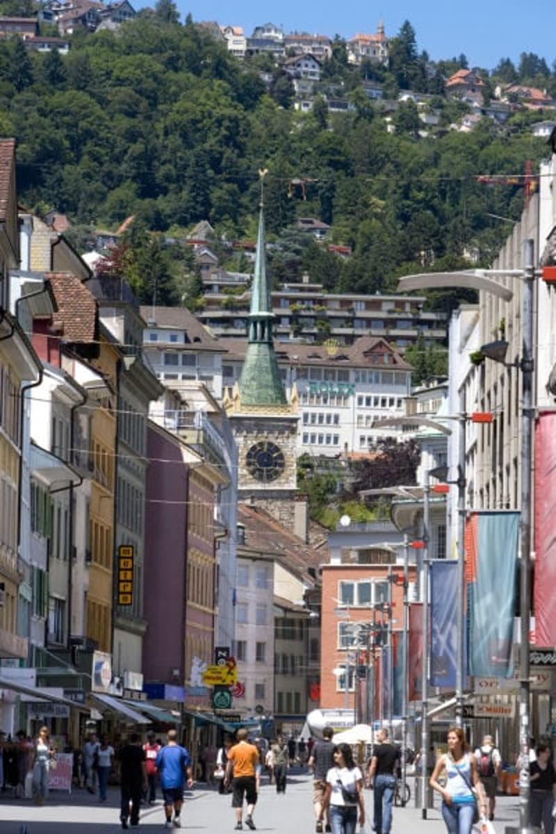 City street with buildings, people walking, clock tower, trees, shops, banners